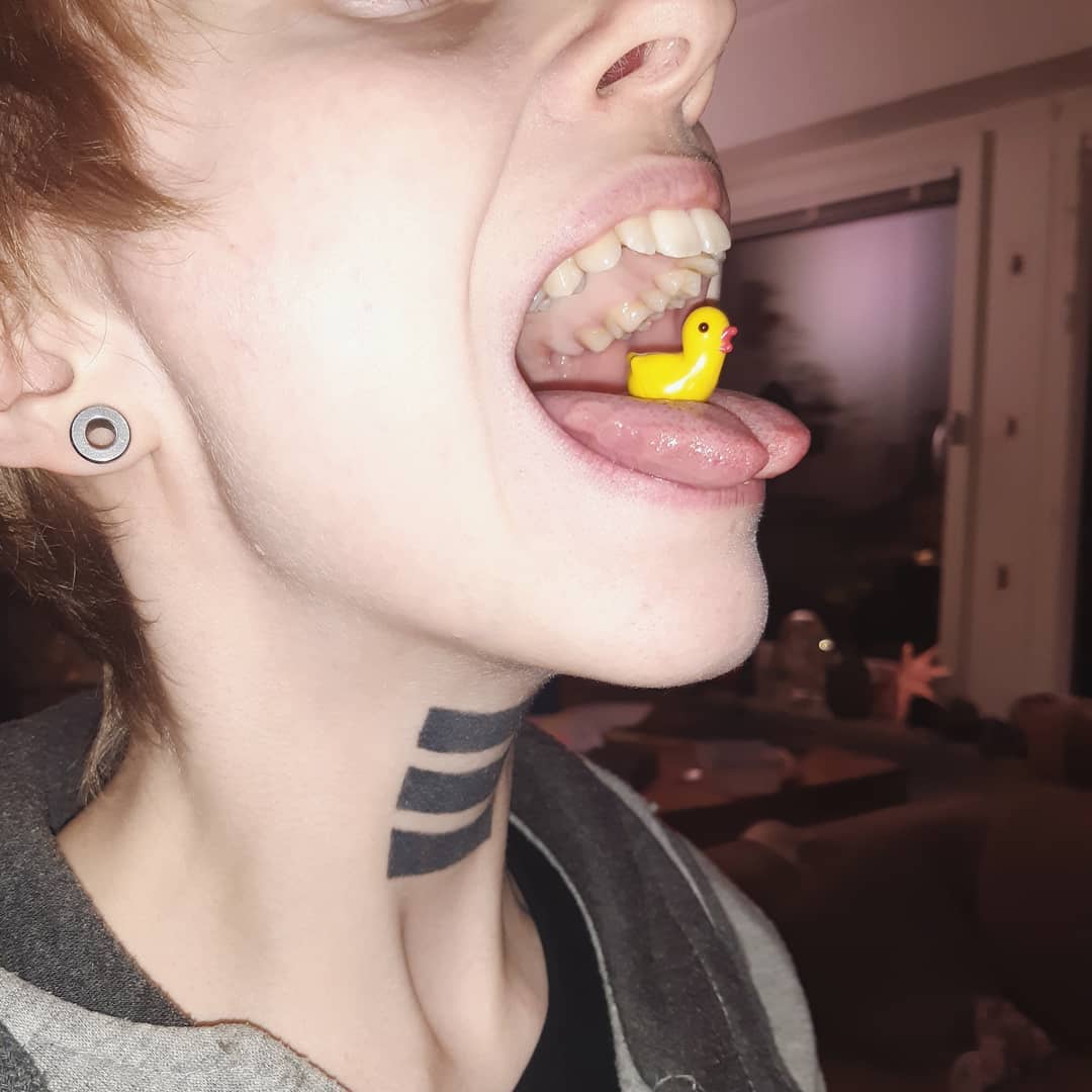 A small, ashtonished porcelain duck on a tongue in an open human mouth. The duck is facing towards the outside.
